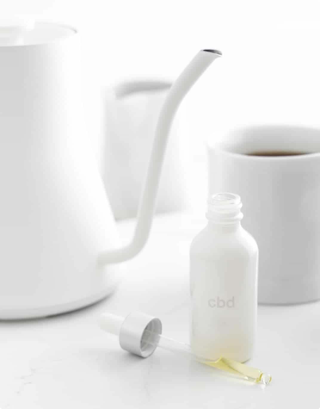 A white teapot, tea cup and open container of CBD oil on a counter.